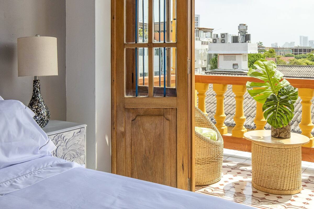A hotel room with a bed, a nightstand with a lamp, a wooden door, and a balcony with wicker furniture and a plant.