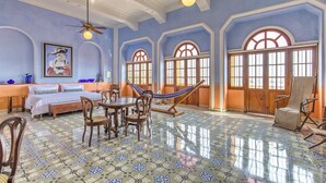 A spacious room with a large bed, a dining table, and chairs. There is a hammock, a ceiling fan, and large windows with arched tops.