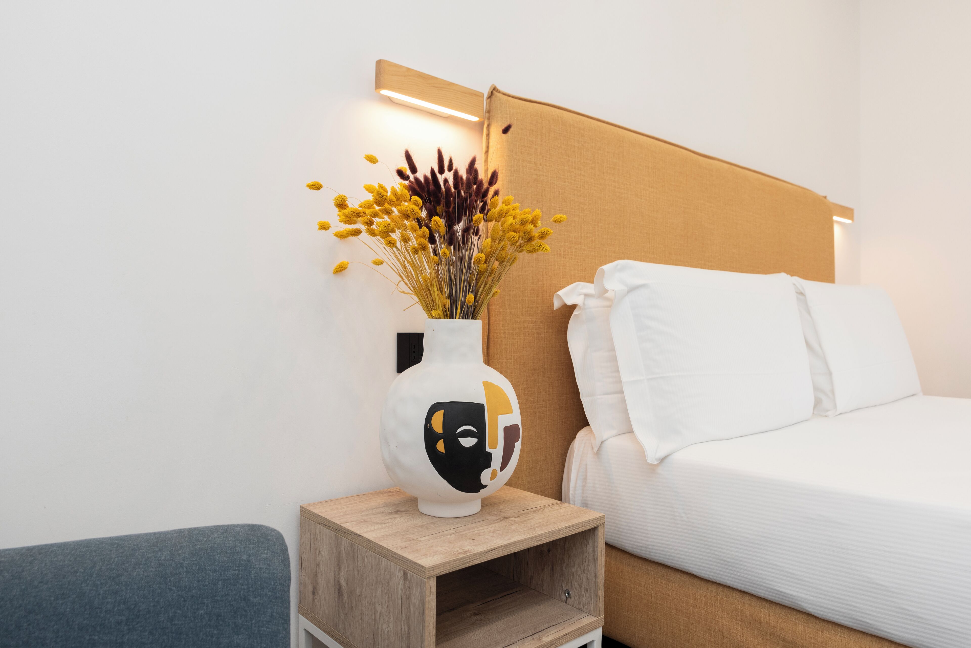 A hotel room with a bed, a bedside table with a vase of flowers, and a wall-mounted light.