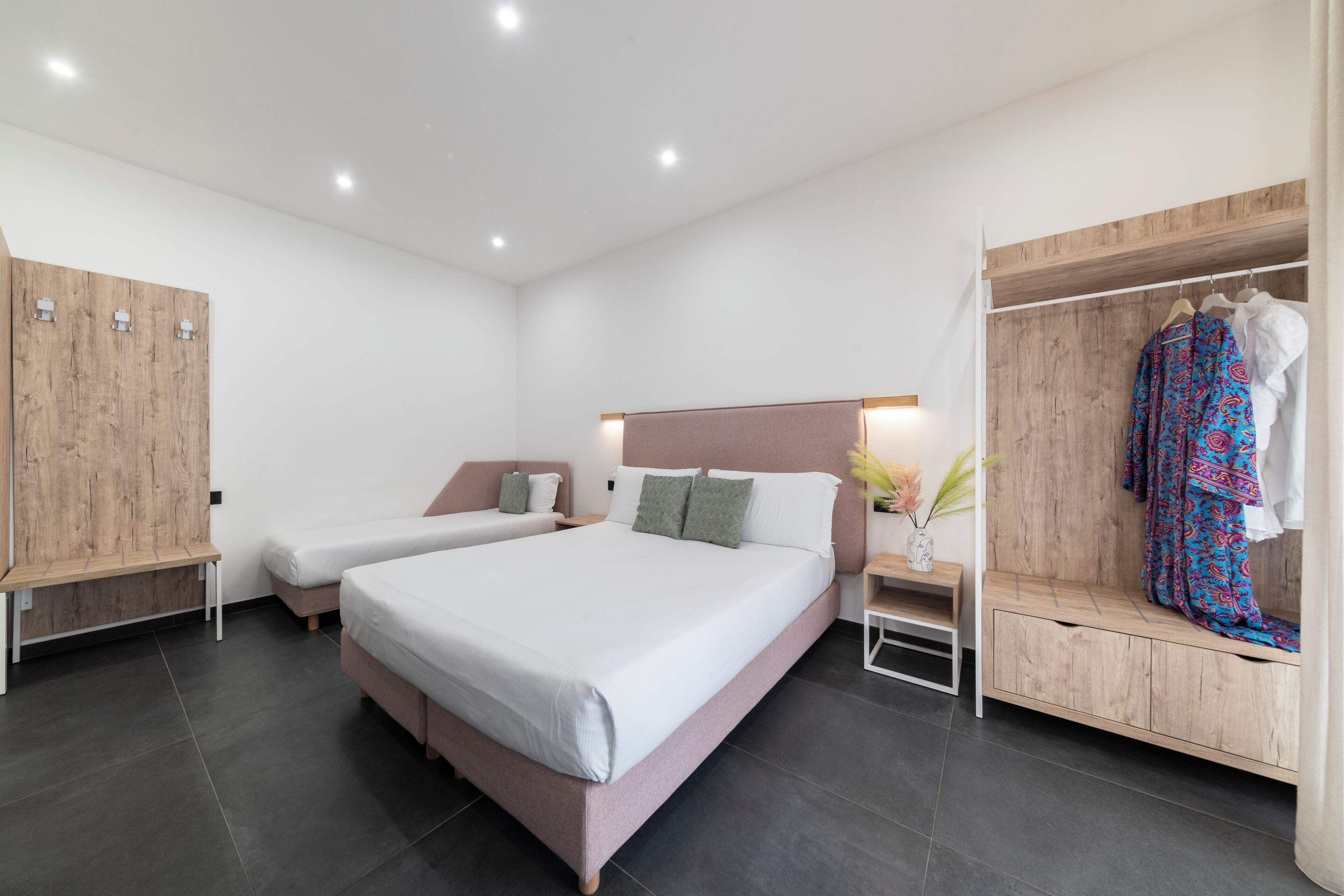 A modern hotel room with a large bed, two smaller beds, wooden wardrobe, and a bench.