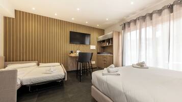 Quarto de hotel com cama, escrivaninha, cadeira, televisão e cozinha compacta.