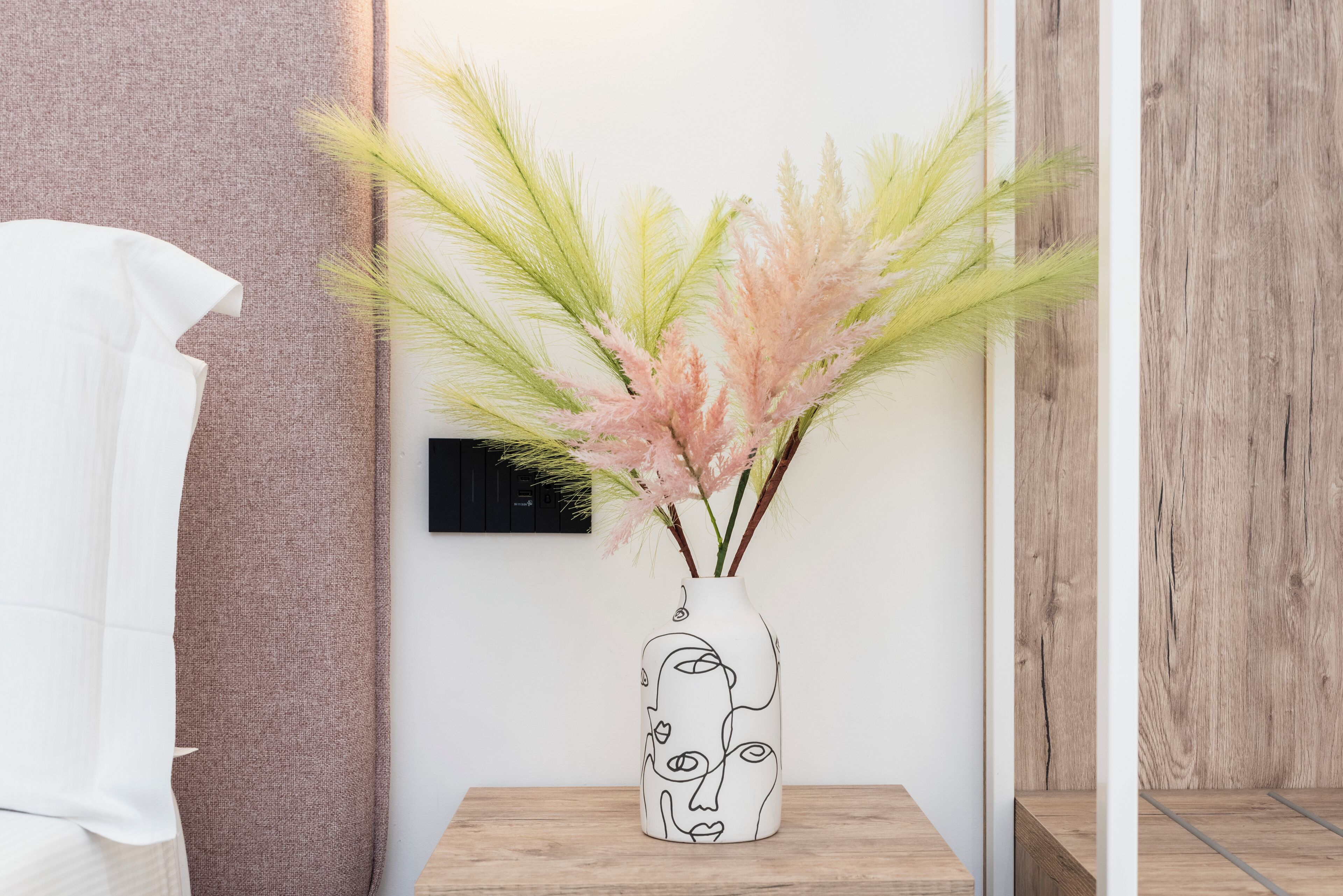 Un jarrón blanco con un diseño facial minimalista sobre una mesa de madera, con hierba de pampa rosa y verde.
