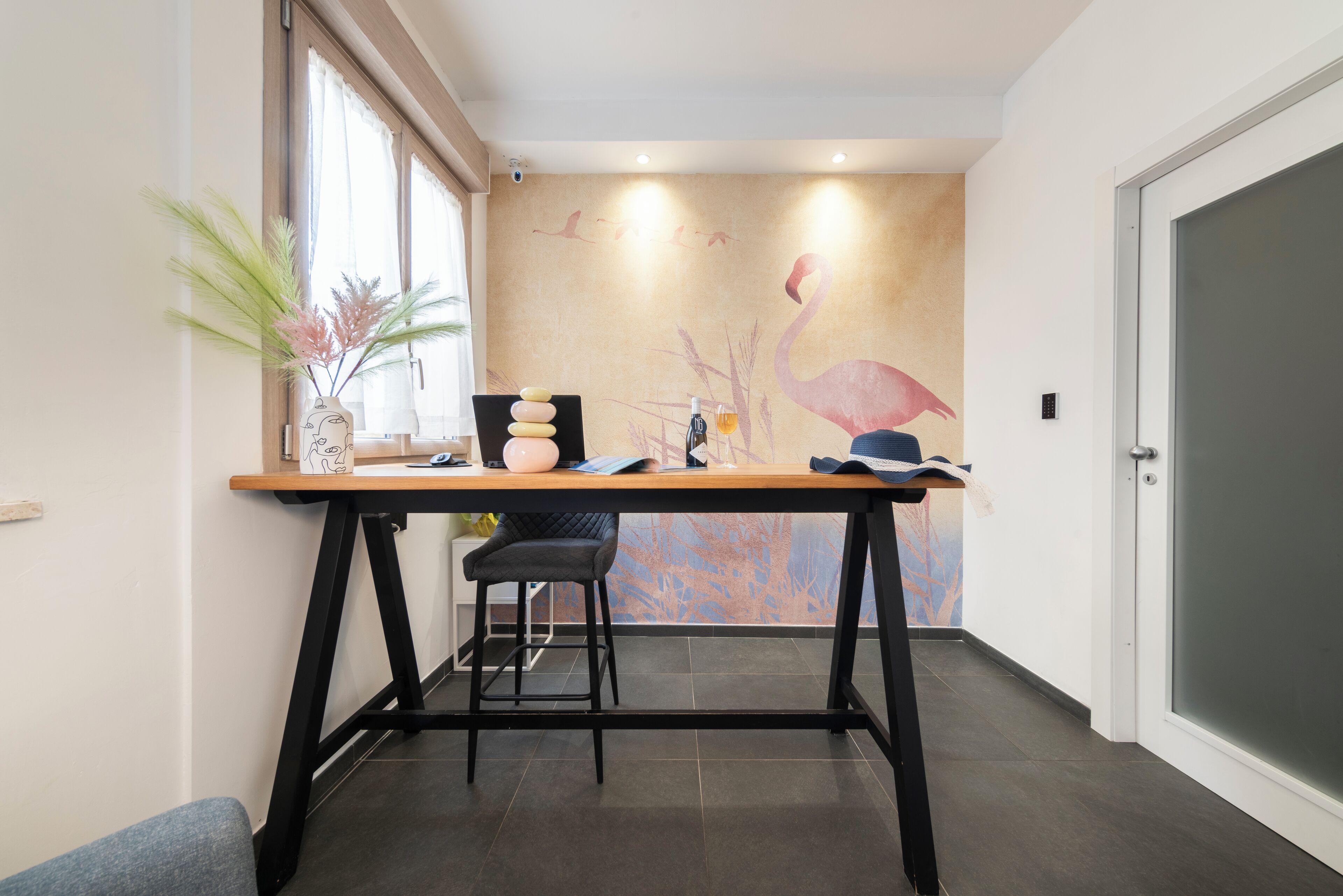 A modern office space with a wooden desk, black metal legs, and a black chair. There is a vase with plants on the left and a wall mural with a flamingo.