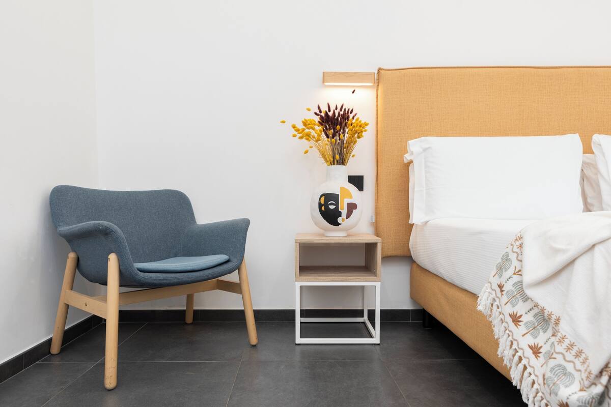 Une chambre moderne avec un lit, une chaise bleue, une petite table sur laquelle est posé un vase de fleurs et un luminaire fixé au mur.