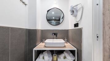 A modern bathroom with a white sink, a round mirror, a towel dispenser, and a toilet paper holder.