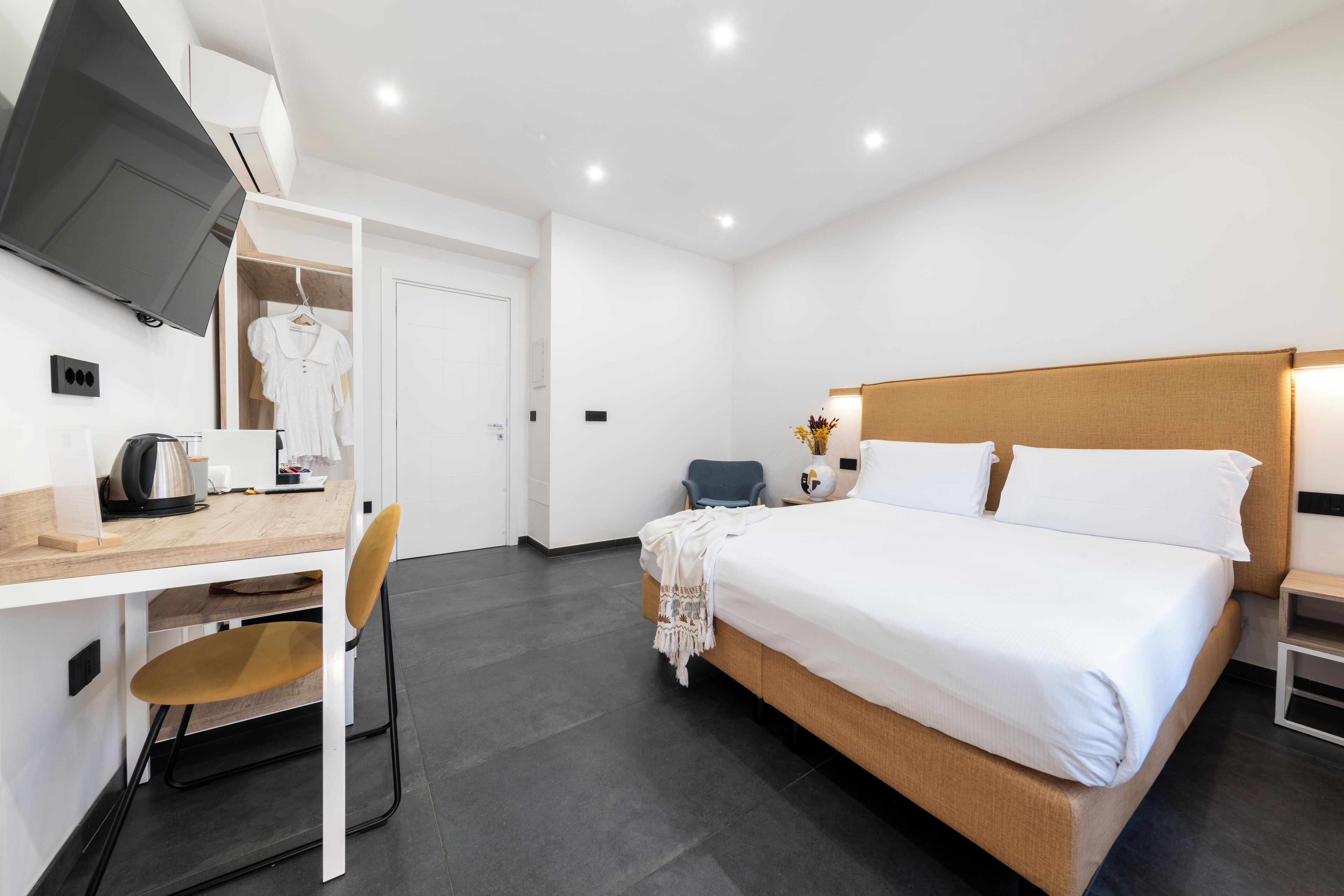 A hotel room with a bed, a desk with a chair, a TV mounted on the wall, and a wardrobe.
