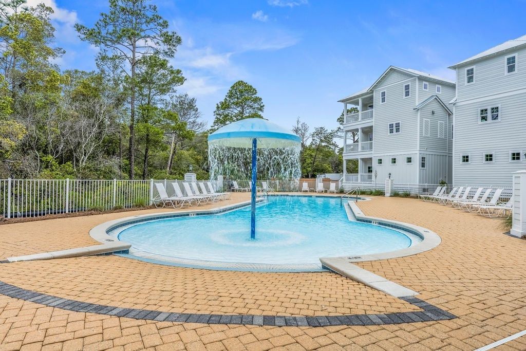 A swimming pool with a fountain, surrounded by lounge chairs and a fenced area with trees.