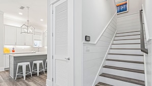 Una cocina moderna con armarios blancos, una isla central con taburetes, una escalera con escalones de madera y un cuadro en la pared.