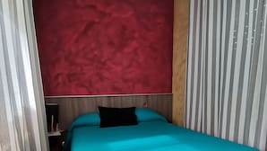 A small, neatly made bed with a teal bedspread and a black pillow, positioned against a red textured wall. - RURAL HOUSE  (Yebra)