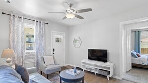 Una sala de estar moderna con un ventilador de techo, un televisor sobre un soporte blanco, un sofá gris y una cortina de ventana estampada.
