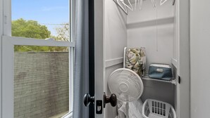 Habitación pequeña con ventana, puerta, ventilador, perchero y un estante con objetos.