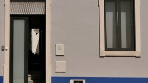 Un bâtiment dont la partie inférieure est bleue, la partie supérieure est blanche, et qui possède une porte avec une grille en treillis métallique, une fenêtre et un petit boîtier électrique.