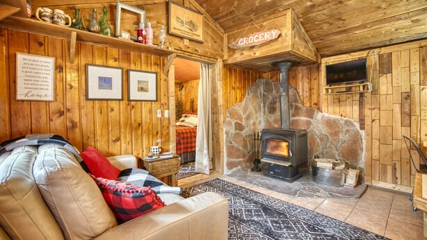 L’intérieur d’un chalet accueillant, avec une cheminée en pierre, un canapé, un mur en bois sur lequel est accroché un panneau indiquant « ÉPICERIE », et un téléviseur fixé au mur.