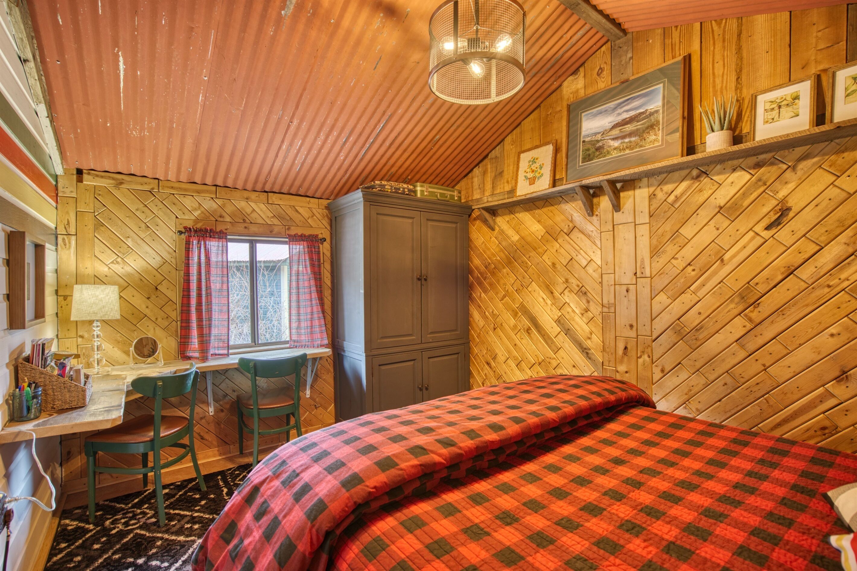 Une pièce chaleureuse avec un mur décoré d’un motif en chevrons en bois, un lit recouvert d’une couverture à carreaux, un bureau avec une chaise et un meuble de rangement.