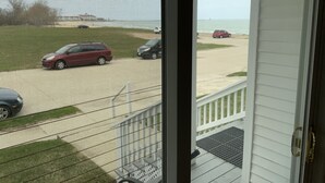 A view through a window with a mesh screen, overlooking a parking area with several cars, a grassy area, and a beach in the distance. - BEACHSIDE GETAWAY IN SHEBOYGAN (Sheboygan)