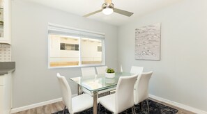 A dining area with a glass table, white chairs, a ceiling fan, and a window with blinds. - Ocean View- One block from ocean & 5 minutes from town! (Del Mar)