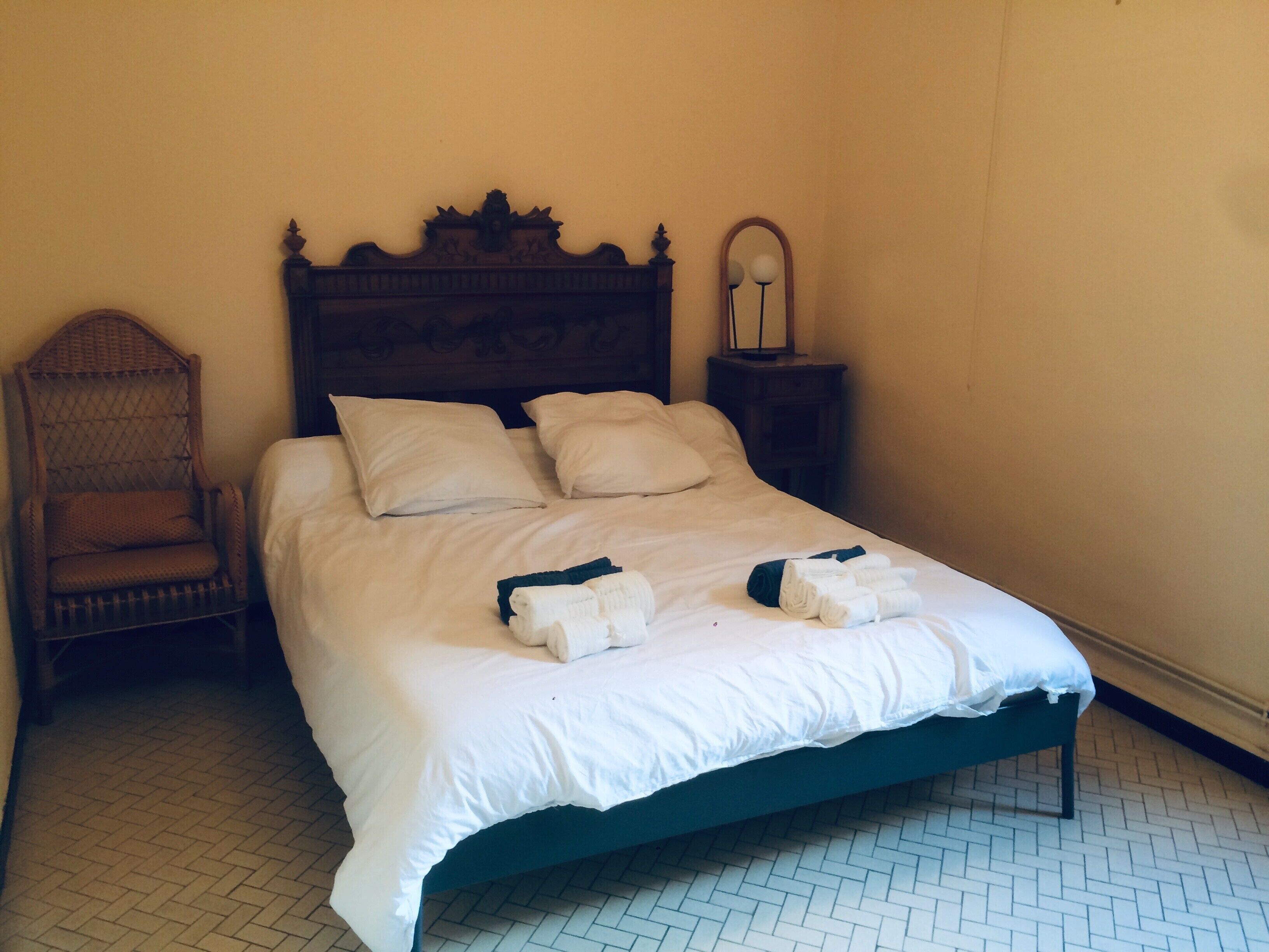 Un lit bien fait, avec du linge de lit blanc et deux serviettes pliées, une tête de lit en bois, une chaise en osier et une petite table de chevet avec un miroir.