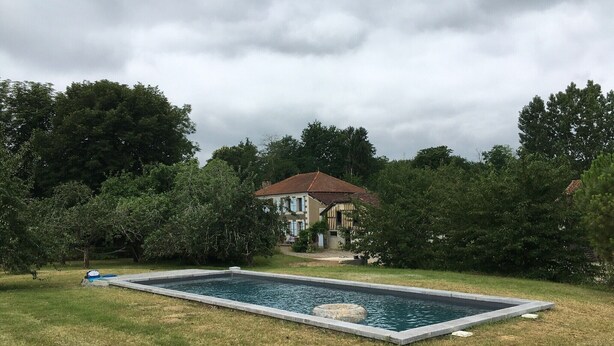Ein Schwimmbecken mit Steinrand, umgeben von Gras und Bäumen, mit einem Haus im Hintergrund.