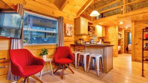 Interior de una cabaña de madera con cocina, dos sillas rojas, una barra y una mesa de comedor.