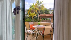 Un balcón con una mesa y sillas, una pequeña planta en maceta y vistas a un jardín con palmeras.