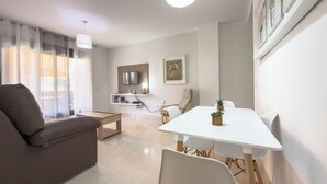 Una sala de estar moderna con un sofá, una mesa de comedor con sillas, un televisor montado en la pared y una lámpara de techo.
