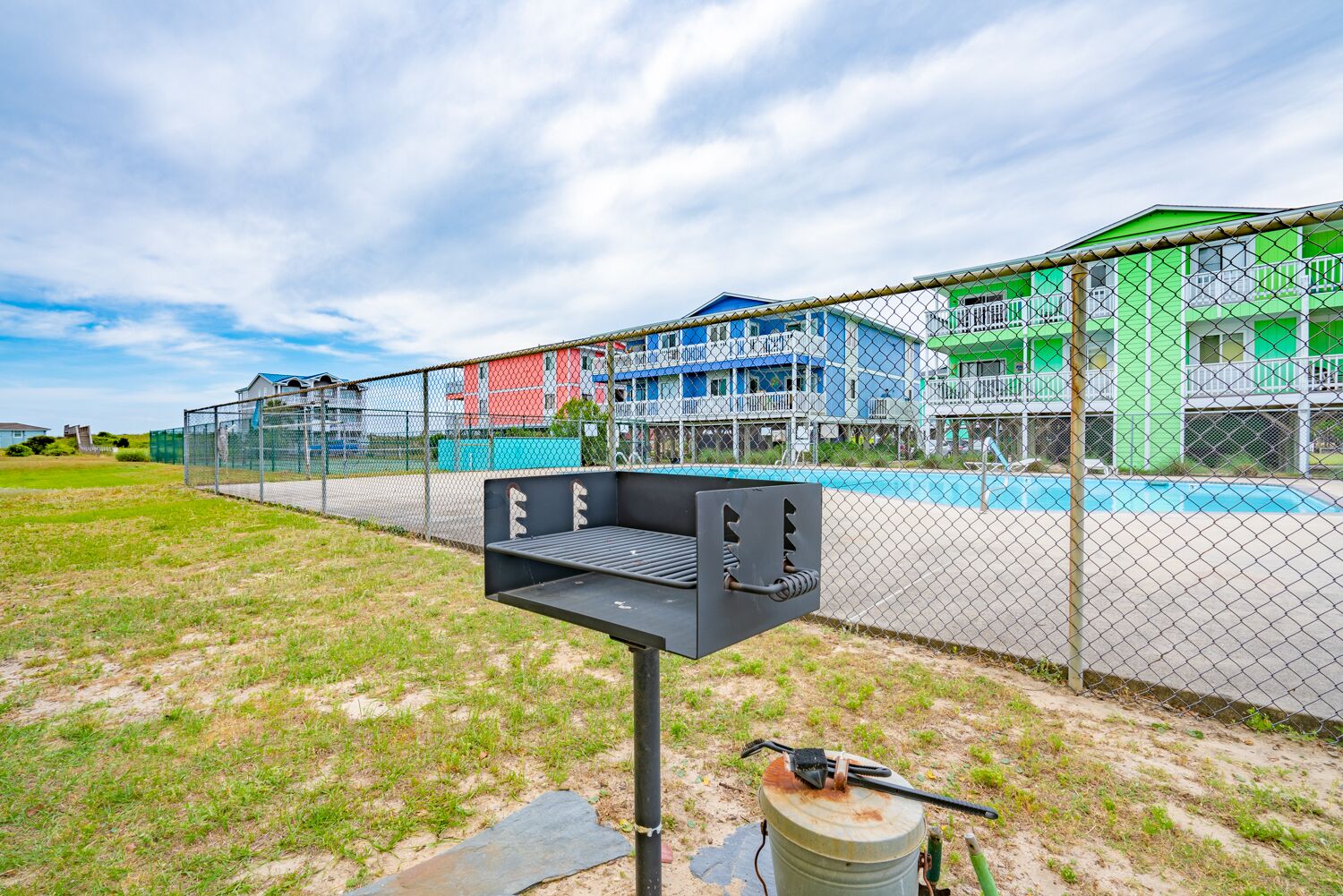 A fenced outdoor area with a metal grill and a barrel, likely for grilling, in front of a building with a pool and a blue and red facade.