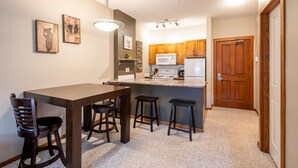 A compact kitchen with wooden cabinets, a microwave, and a coffee maker. There is a dining table with four stools and two wall-mounted light fixtures. - Ski in, Ski out Fireside Retreat  (Sun Peaks)