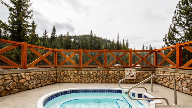 A hot tub with a stone wall and wooden railing, surrounded by a forested area. - Ski in, Ski out Fireside Retreat  (Sun Peaks)