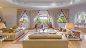 A spacious living room with a large beige sofa, two armchairs, a coffee table, and a dining area with a table and chairs. The room features large windows with curtains and a view of greenery outside. - Palatial Villa in Hacienda Las Chapas, Marbella (Marbella)