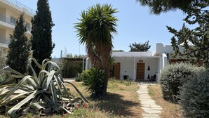 Un sendero conduce a un edificio blanco con puerta de madera, rodeado de plantas y árboles.