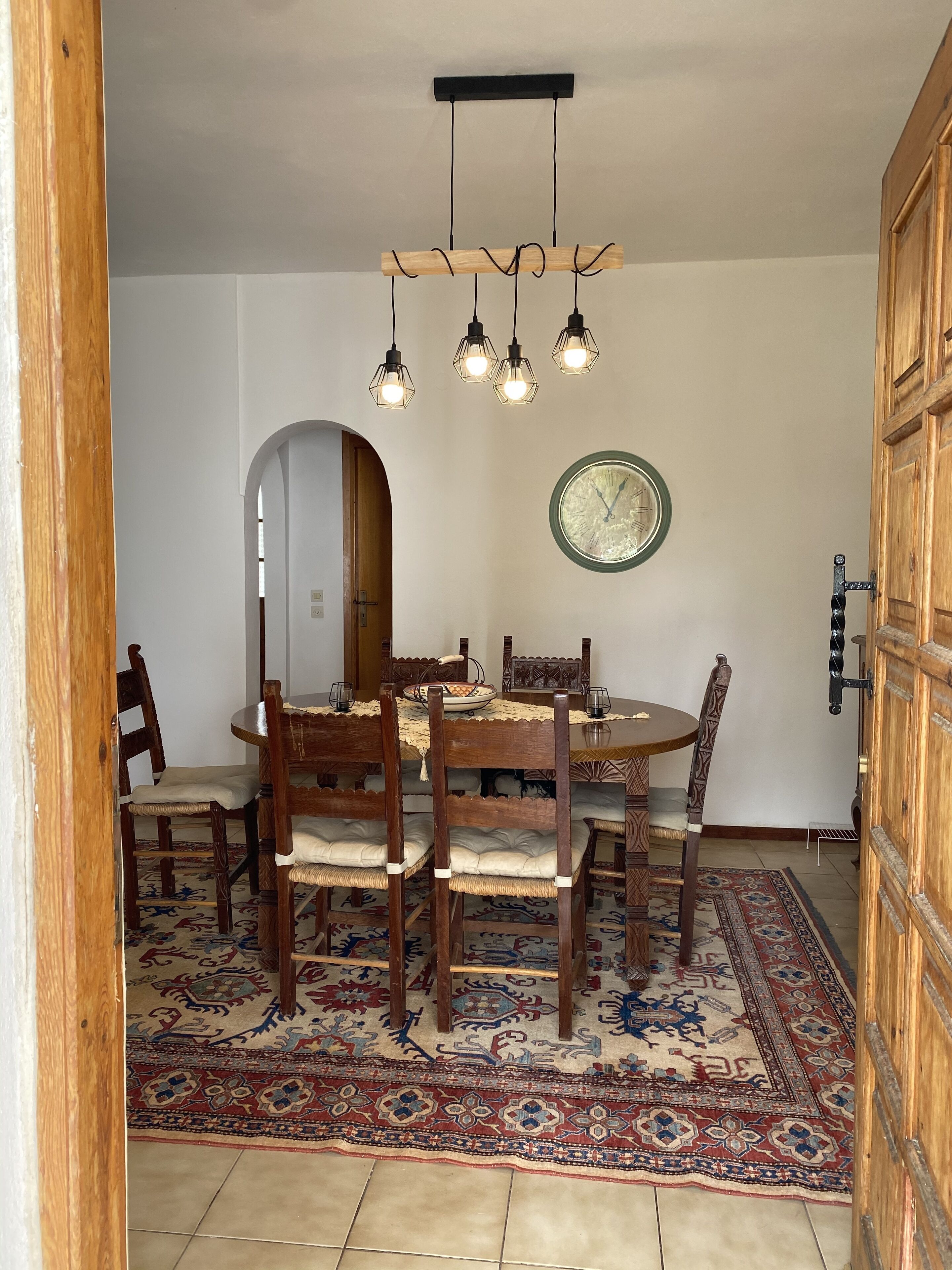 Zona pranzo con tavolo rotondo, sedie in legno, tappeto decorato, orologio a muro e porta in legno.