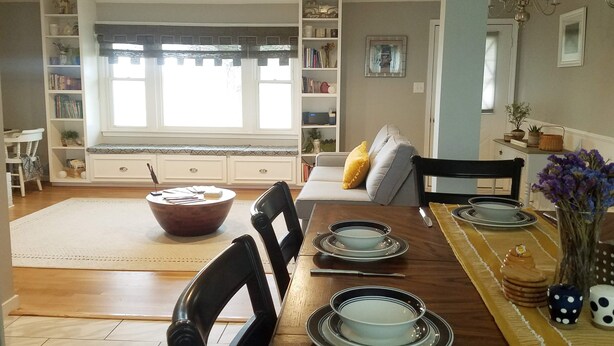 Un comedor con una mesa de madera preparada para cuatro personas, rodeado de estanterías y una ventana con cortinas.