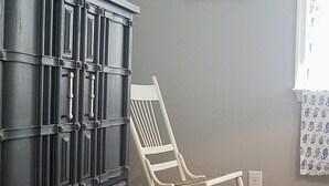 Una habitación con un aparador de madera oscura, una mecedora blanca, un cuadro enmarcado en la pared y una ventana con cortina estampada.