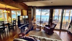A living room with a TV, dining table, and chairs, overlooking a deck with a view of the sea. - Pine Cottage - Beautiful direct lakefront cottage located in Sodus, NY, with full amenities. (Sodus)
