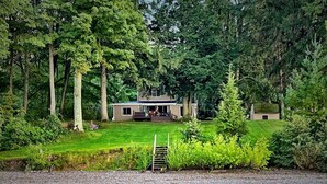 A lakeside cabin with a deck, surrounded by dense forest and a gravel path leading to the water. - Pine Cottage - Beautiful direct lakefront cottage located in Sodus, NY, with full amenities. (Sodus)