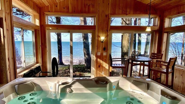 A wooden cabin with a hot tub and a table and chairs by a large window overlooking a body of water. - Pine Cottage - Beautiful direct lakefront cottage located in Sodus, NY, with full amenities. (Sodus)