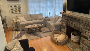 A living room with a beige sofa set, a coffee table, a dog bed, a fireplace, and a TV mounted on the wall. - Brand new year round house with easy access to to everywhere in Ogunquit (Ogunquit)