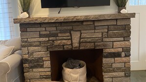 A living room with a stone fireplace, a television mounted above it, and a dog bed with a dog lying inside. - Brand new year round house with easy access to to everywhere in Ogunquit (Ogunquit)