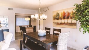 Un coin repas avec une table en bois foncé, des chaises blanches, un lustre et un tableau représentant des arbres accroché au mur.