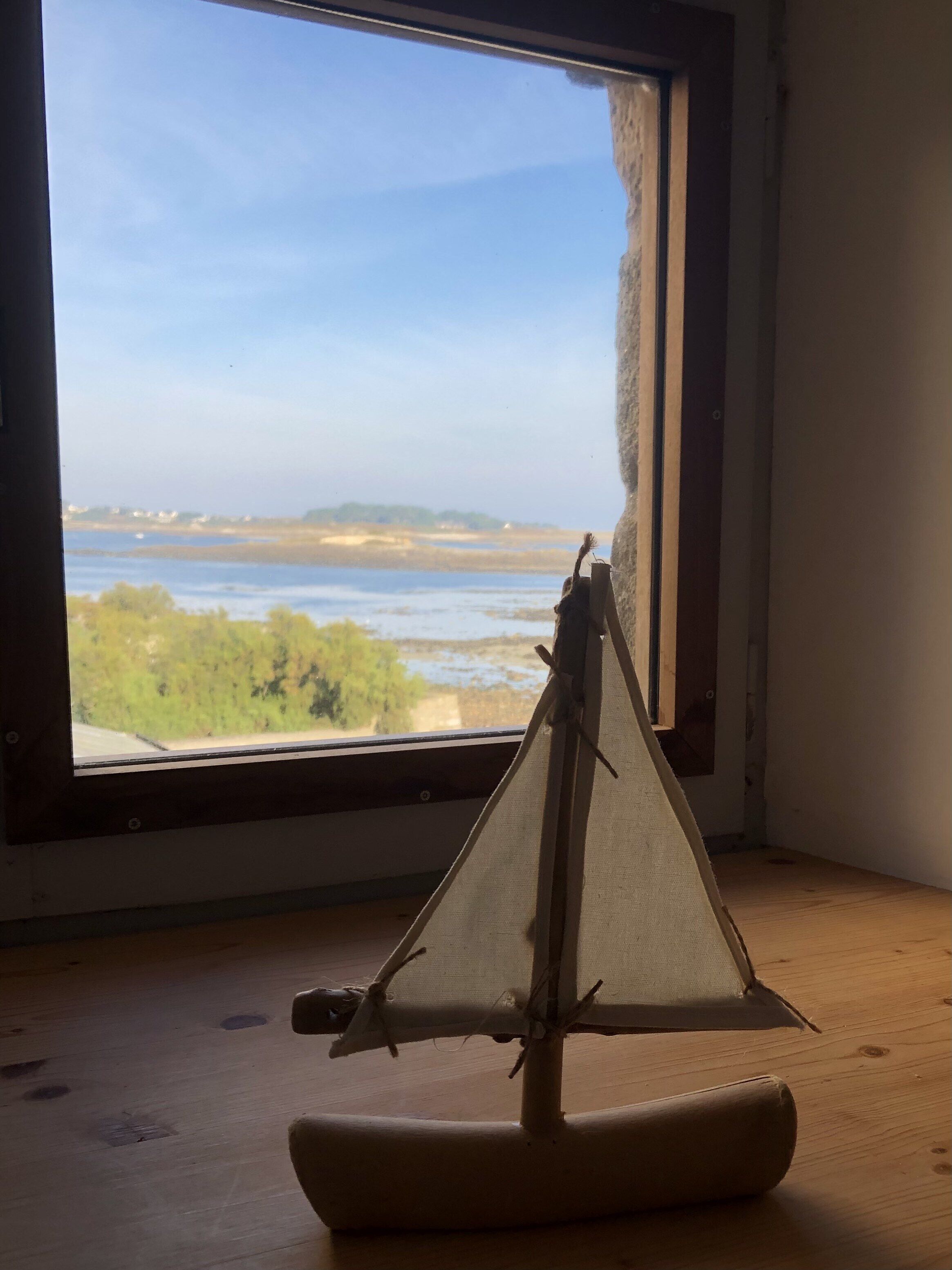 A model sailboat on a wooden surface near a window with a view of a body of water and distant land.