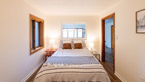 Une chambre avec un grand lit, deux tables de chevet avec des lampes, une fenêtre avec des stores et un tableau encadré au mur.