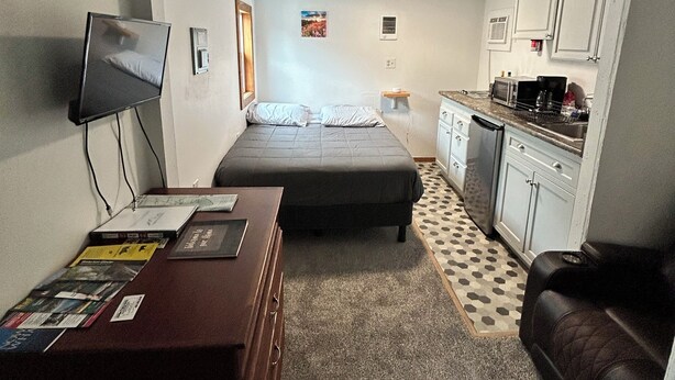 A compact hotel room with a bed, a desk with a computer, a kitchenette, and a small seating area. - Teakettle Kabin Tiny Home (Columbia Falls)
