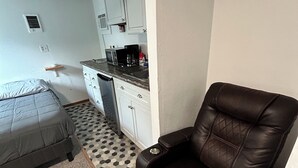 A compact room with a bed, a brown leather chair, a kitchenette, and a patterned rug. - Teakettle Kabin Tiny Home (Columbia Falls)