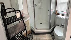 A compact bathroom with a glass shower enclosure, a toilet, a sink, and a towel rack. - Teakettle Kabin Tiny Home (Columbia Falls)