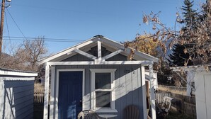 A small, single-story building with a blue door and a window, a patio with two chairs and a table, and a fenced yard. - Teakettle Kabin Tiny Home (Columbia Falls)