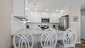 A modern kitchen with white cabinetry, a stainless steel refrigerator, a microwave, and a coffee maker. There are three white chairs with patterned cushions. - Almost Phamily! - 2 bedroom cottage in Atlantic Beach (Atlantic Beach)
