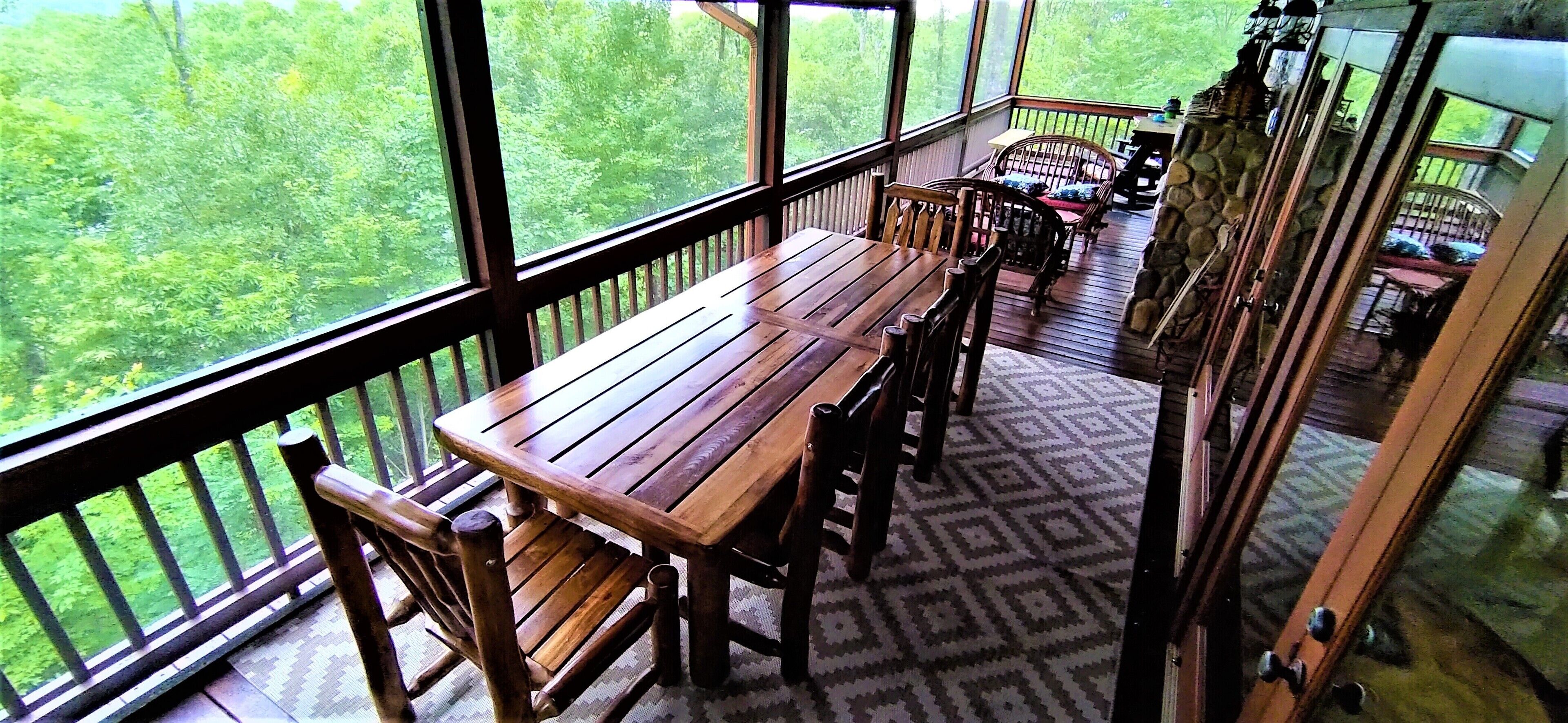 A wooden deck with a table and chairs, large windows offering a view of a forest, and a stone wall on one side.