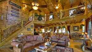 Ein geräumiges Blockhaus-Interieur mit hohen Decken, Holzdielen und einer zentralen Sitzecke mit zwei Ledersofas und einem Couchtisch.