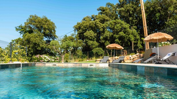Una piscina cristalina con sillas de descanso y sombrillas, rodeada de árboles y un muro de piedra.