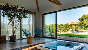 Una habitación moderna con suelo de madera, un gran ventanal corredero que da a un jardín, un jacuzzi y dos sillas azules.